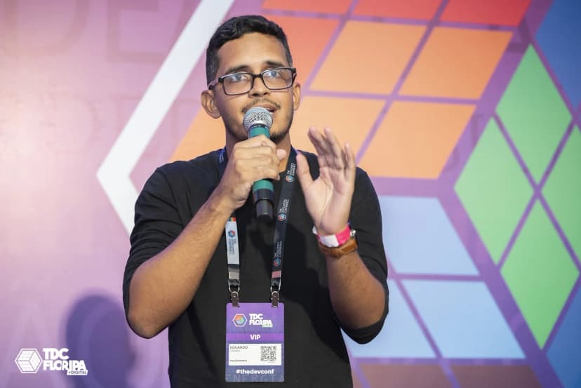 Eduardo Corrêa falando em palco durante evento de tecnologia, com fundo colorido e banner do TDC Floripa, usando blusa preta e óculos, segurando microfone.